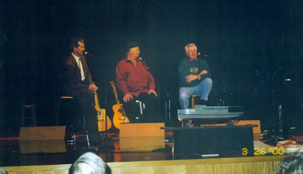 Three men sit on a stage with microphones, guitars, and audio equipment while audience members watch.