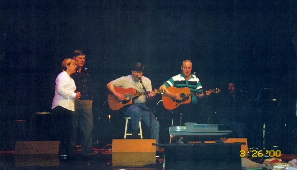 Five people perform on a dimly lit stage, including two acoustic guitarists and a singer near a microphone.