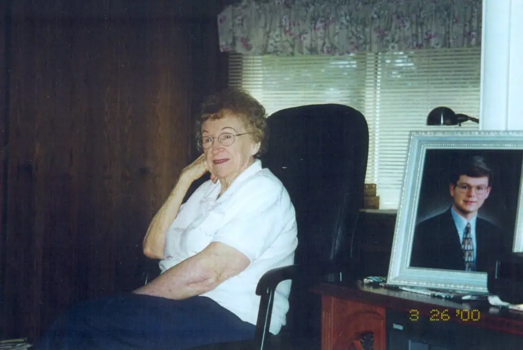An older woman sits in an office beside a framed portrait and a desk.