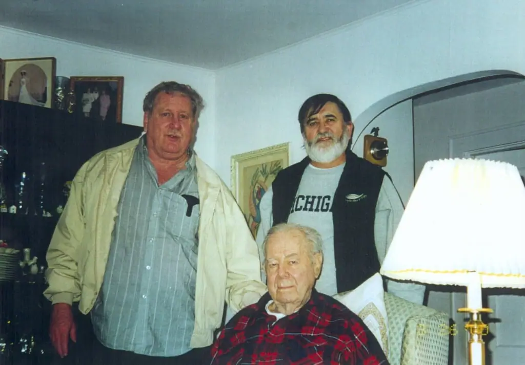 Three men pose for a photograph in a furnished living room, with one seated and two standing behind him.