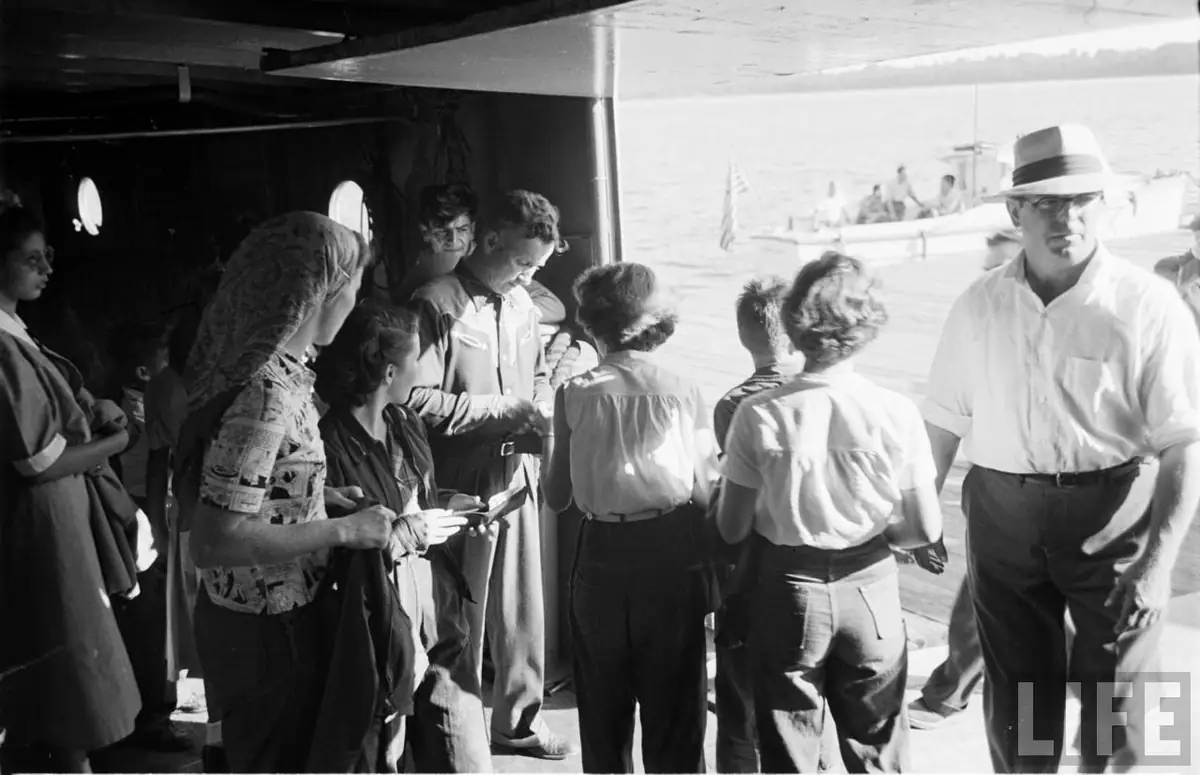 A group gathers on a ferry deck beside a man wearing a fedora, with another boat and shoreline visible beyond.