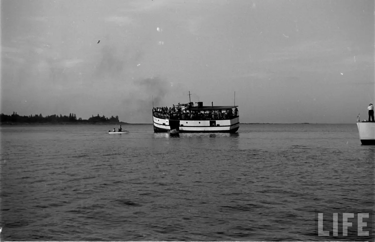 Black-and-white photograph of a crowded ferry with smaller boats on open water.