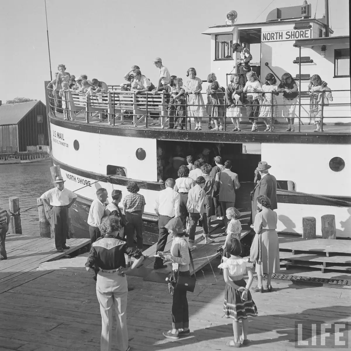 Crowded ferry beside a wooden pier, with passengers boarding and gathered on the upper deck.
