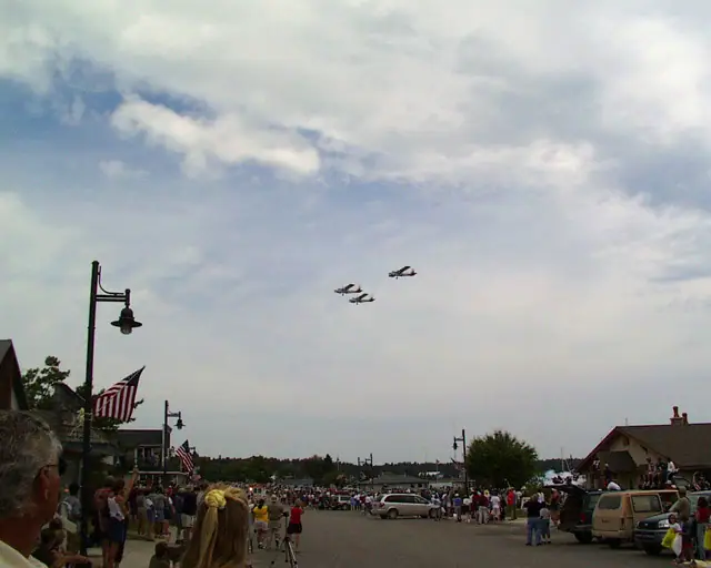 Four small airplanes fly in formation above a crowded street lined with flags and spectators.
