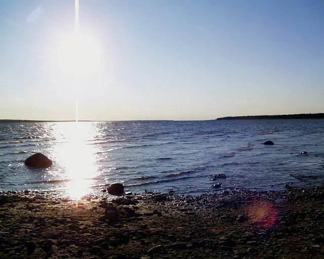 Low sun glitters across calm water beyond a rocky shoreline, with a dark distant shore on the horizon.