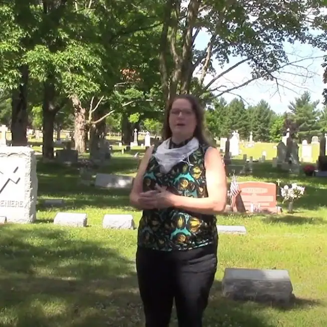 A woman stands among gravestones beneath trees in a cemetery.