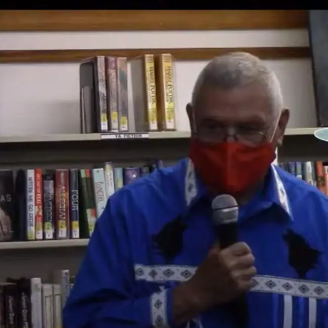 Masked man holding a microphone in front of a bookshelf.