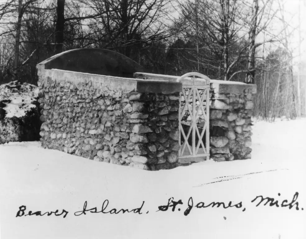 Snow-covered stone enclosure with an arched lattice gate among leafless trees.