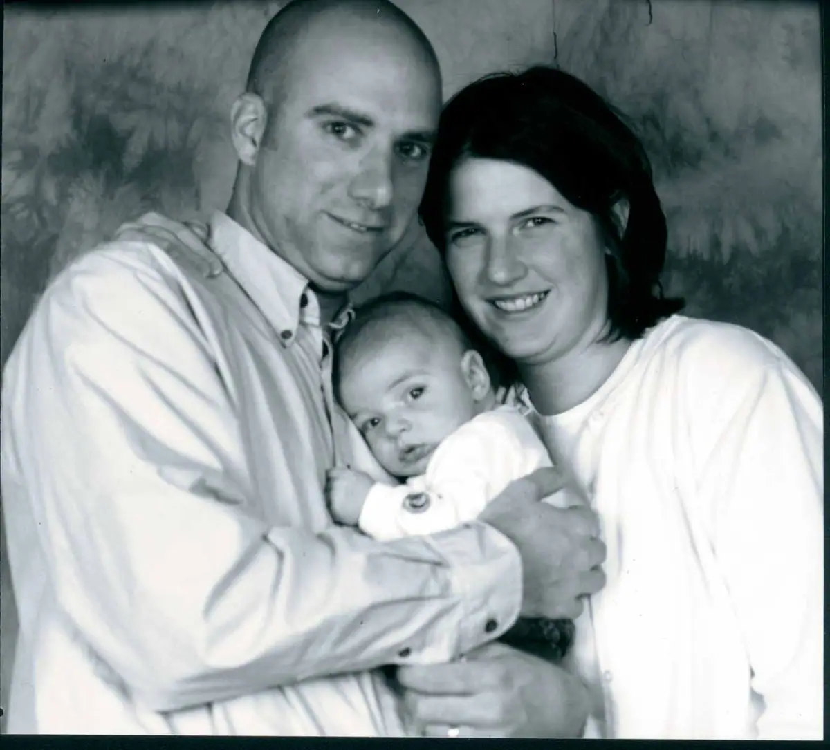 Black-and-white portrait of two adults holding a baby against a mottled backdrop.