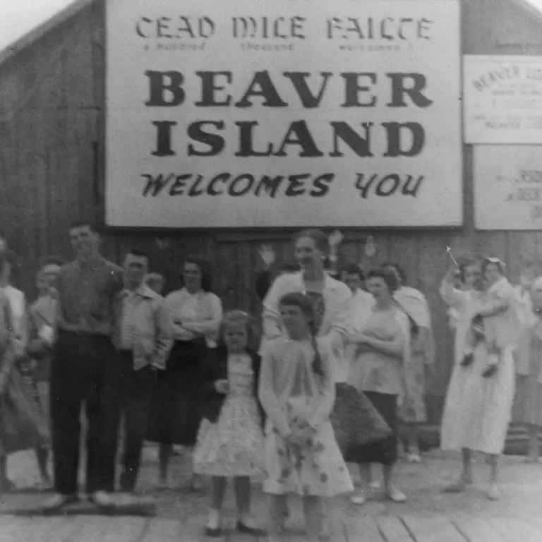 A group of adults and children poses beneath a large bilingual welcome sign outside a wooden building.