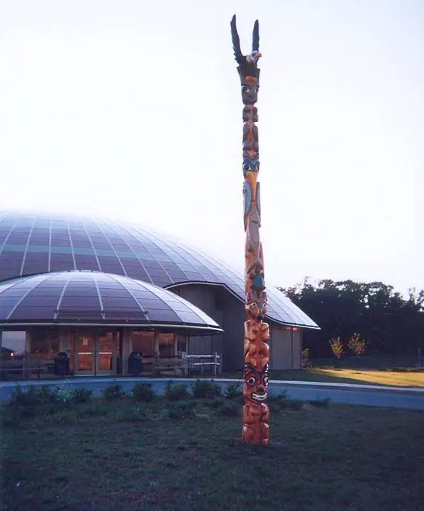 A tall carved totem pole stands on a lawn before a modern building with curved glass-domed roofs.