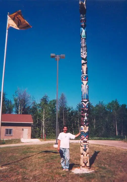 A man stands beside a tall painted and carved wooden totem pole.