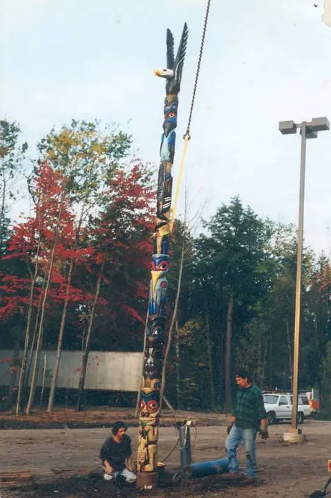 Two people work beside a tall, brightly painted carved pole suspended from a cable outdoors.