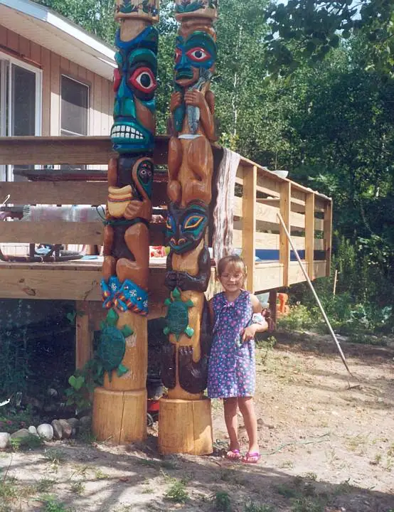 A child stands beside two brightly painted wooden totem poles outside a raised wooden structure.