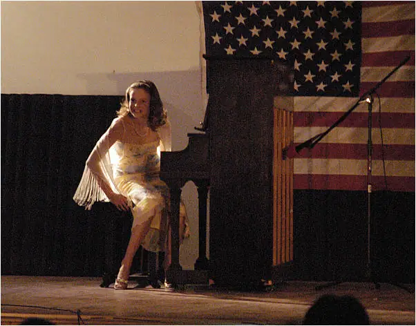 A woman in a light dress sits beside a grand piano on a stage beneath a large American flag.