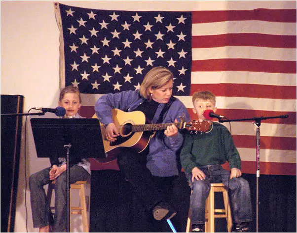 A woman and two children perform with a guitar and microphones beneath a large American flag.