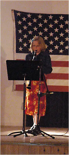 A woman plays a clarinet at a microphone on a stage before a large American flag.