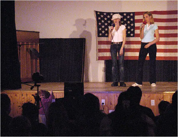 Two women holding microphones perform on a stage before an audience, with an American flag behind them.