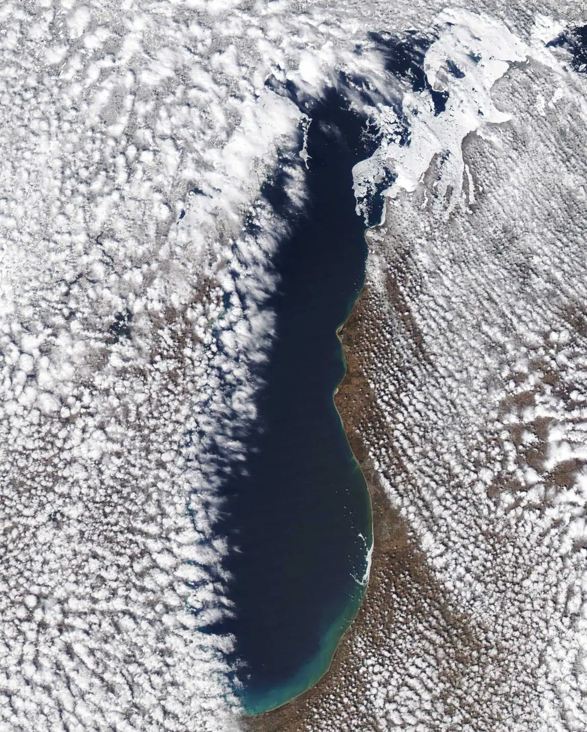 Aerial view of a dark waterway bordered by snow, ice, forest, and dense cloud cover.