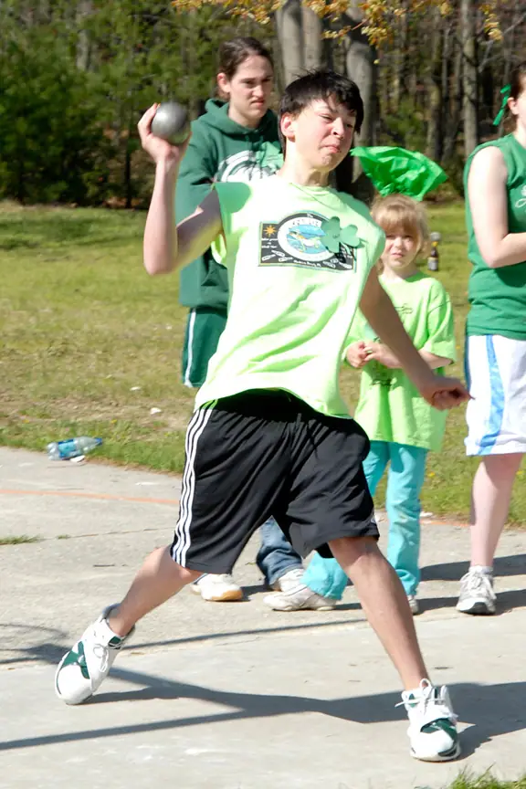 Young athlete lunging forward while throwing a shot put ball, with spectators and teammates behind.