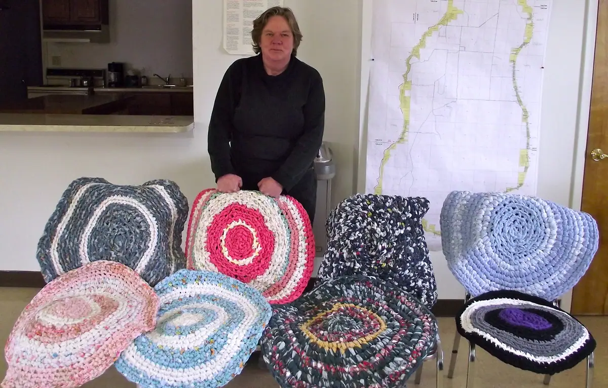 A woman stands behind colorful circular rag rugs arranged on chairs in a room with a wall map.
