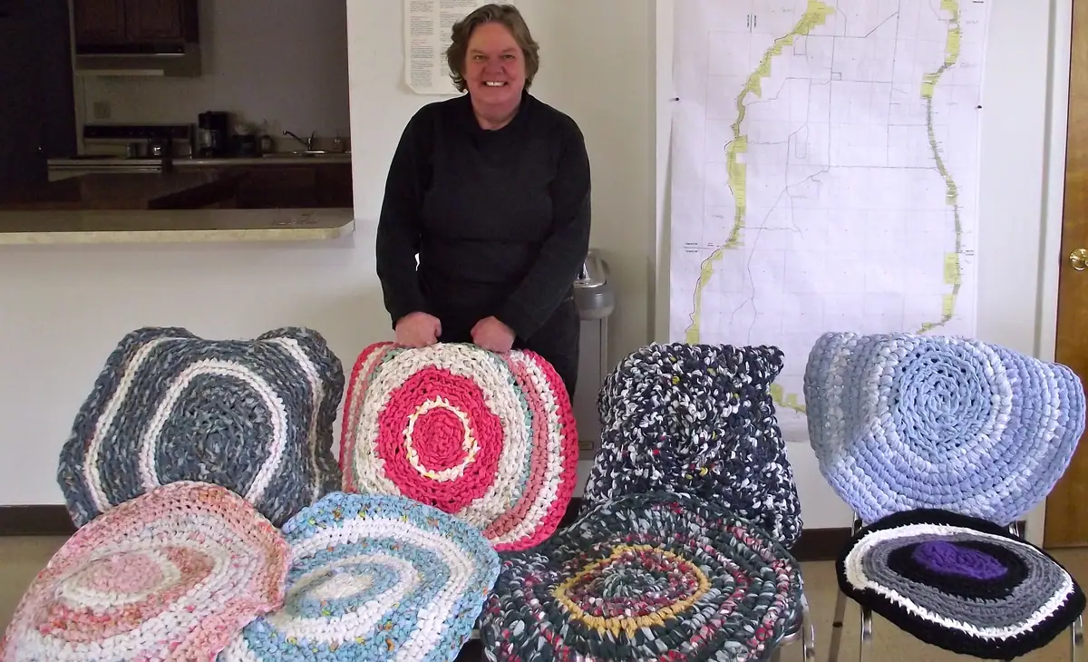 Smiling woman displays colorful circular rag rugs arranged on chairs in an indoor room.