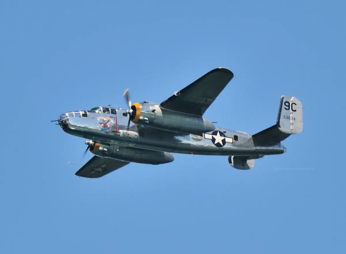 A silver twin-engine propeller aircraft flies against a clear blue sky.