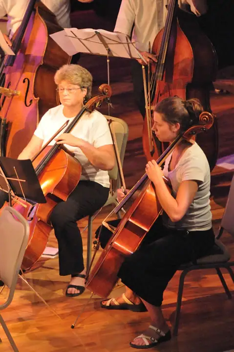 Two seated musicians play cellos while other string players and sheet music appear behind them.