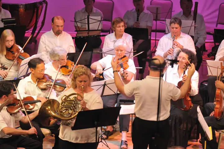 Conductor leading an orchestra of seated musicians on a brightly lit stage.