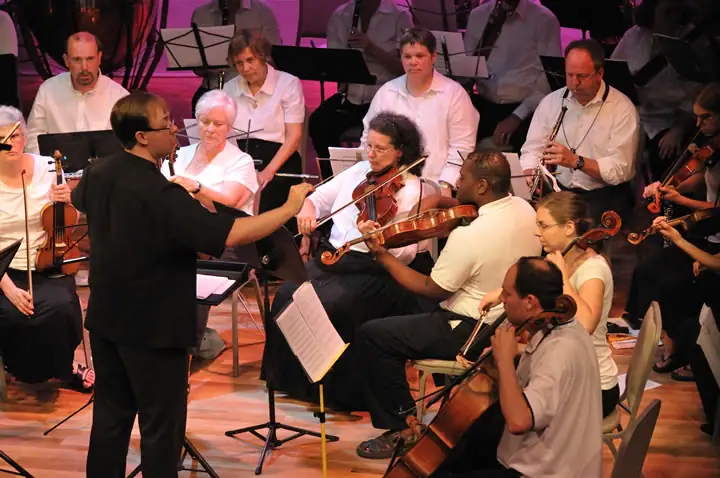 A conductor leads a seated string ensemble performing on a warmly lit stage.
