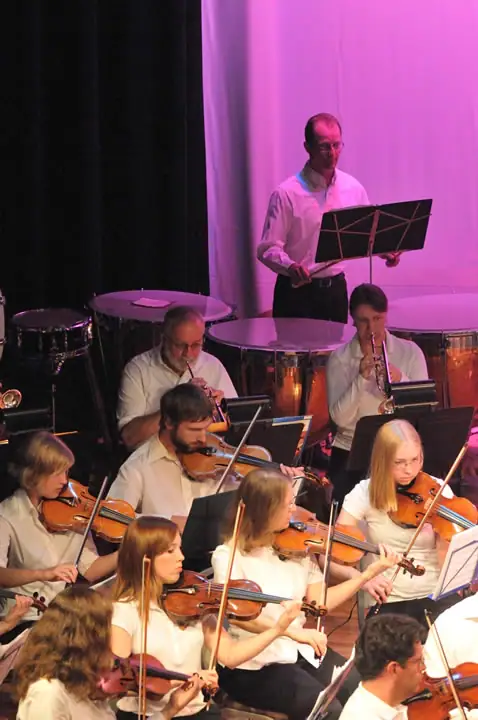 Musicians perform with violins, trumpets, and percussion under pink stage lighting.