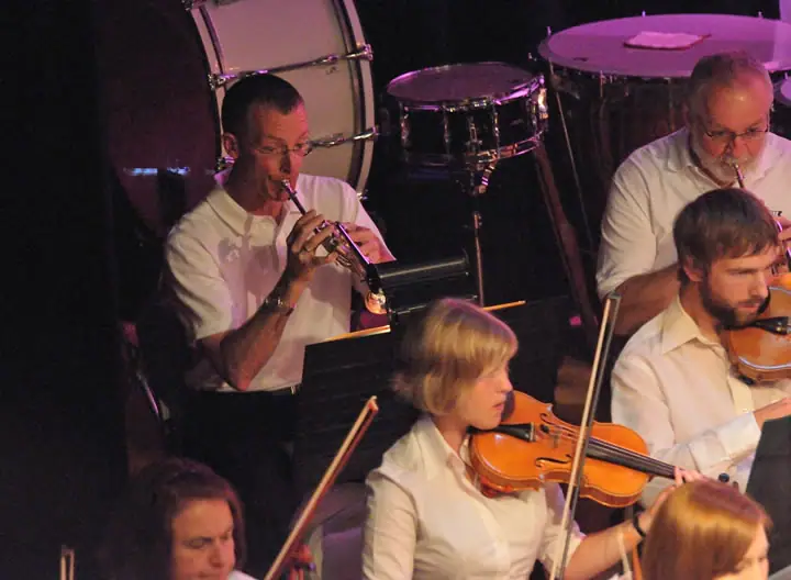 Musicians perform with a trumpet, violins, and percussion instruments under colored stage lighting.