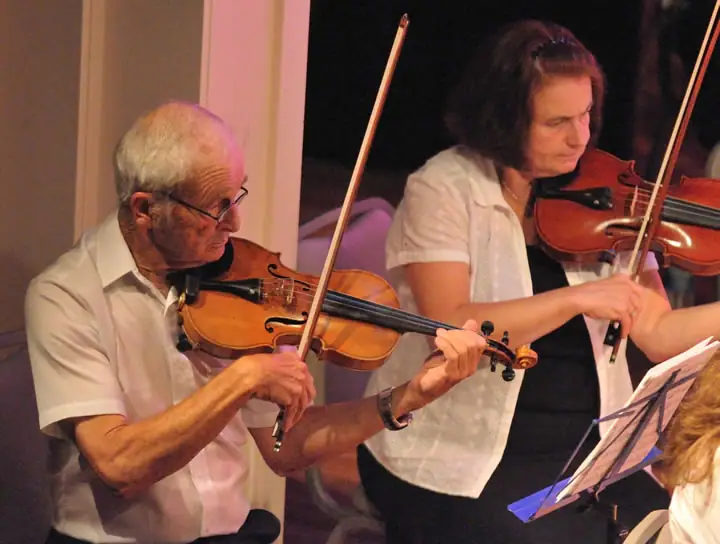 Two violinists perform indoors, with sheet music visible at the lower right.