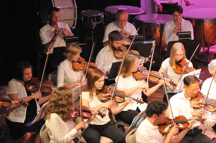 Musicians in white shirts play violins and other instruments on a brightly lit stage.