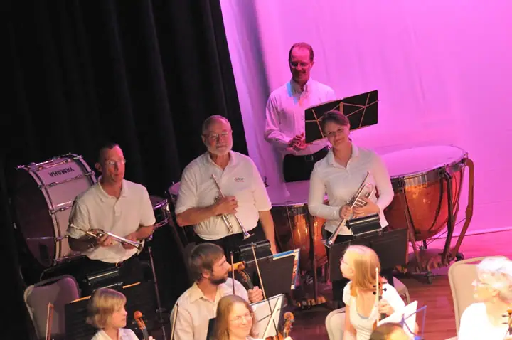 Musicians perform on a brightly lit stage with brass, string, and percussion instruments.
