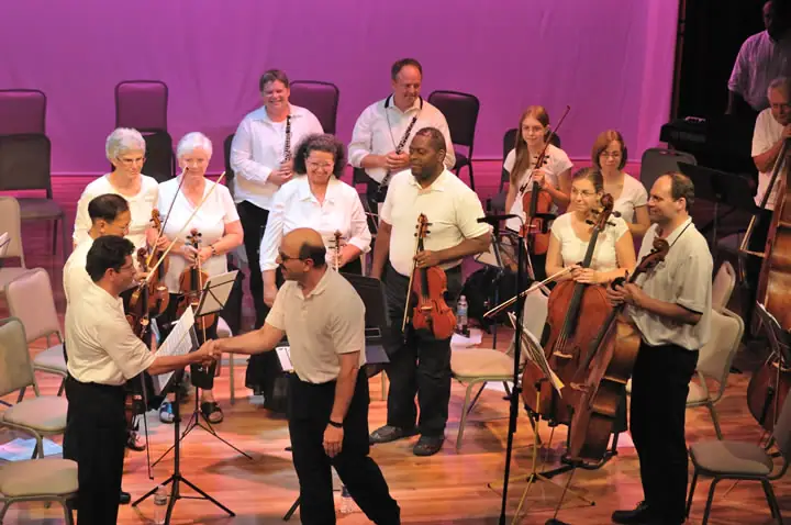 Musicians holding violins, cellos, woodwinds, and a double bass gather on a brightly lit stage.