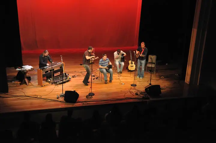 Five musicians perform on a stage before a seated audience.