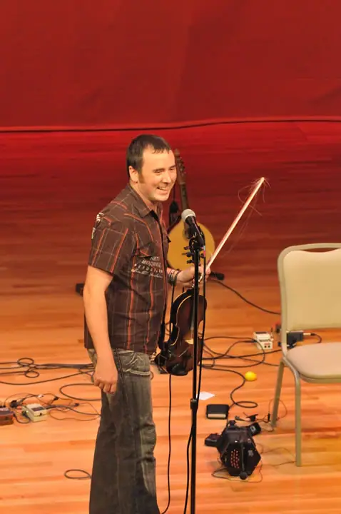 A musician stands with a violin and bow beside a microphone on a stage cluttered with cables and equipment.
