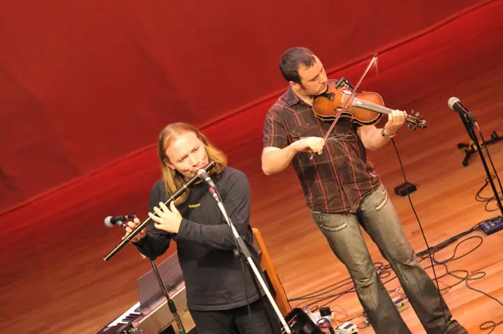 Two musicians perform onstage, one playing a flute and the other a violin.