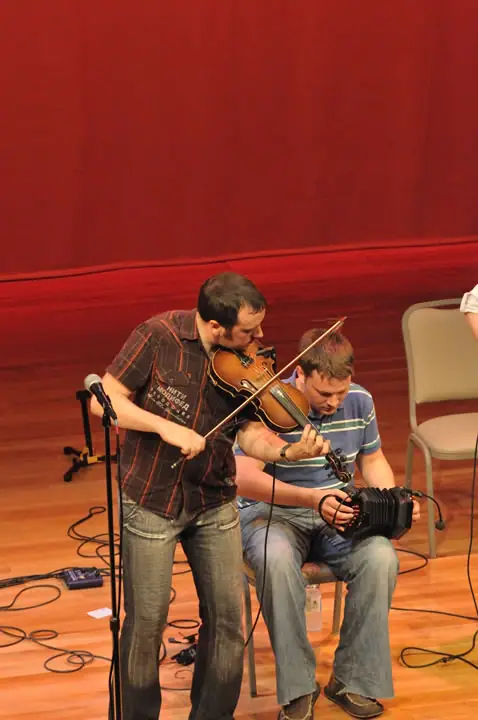 Two men perform on a stage, one playing violin and the other accordion.