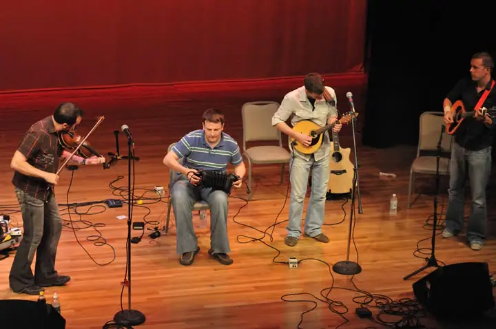 Four musicians perform on a stage with string instruments and an accordion before a red curtain.
