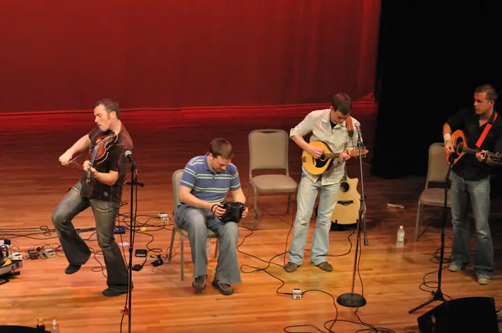 Four people appear on a wooden stage, with three playing string instruments beneath a red curtain.