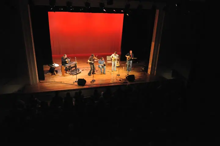 Five musicians perform on a lit stage before a seated audience.