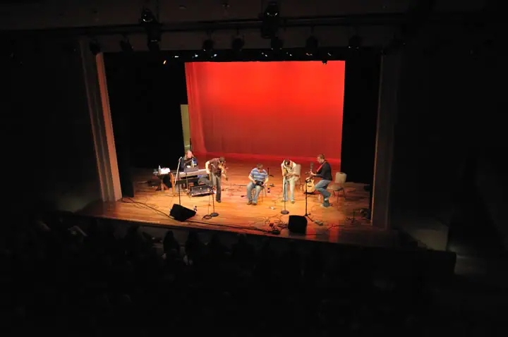 Musicians perform on a lit stage before a seated audience, with a large red backdrop behind them.