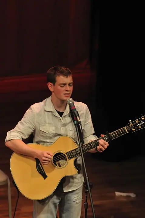 Musician singing into a microphone while playing an acoustic guitar on a dimly lit stage.