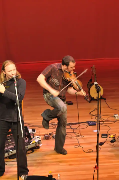 Two musicians perform on a wooden stage, with a violin, stringed instrument, microphones, and audio cables visible.