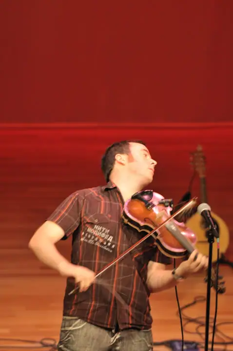 A musician plays a violin beside a microphone on a stage with red curtains.