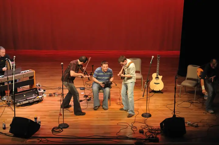 Musicians perform on a stage with guitars, a violin, a keyboard, microphones, and sound equipment.