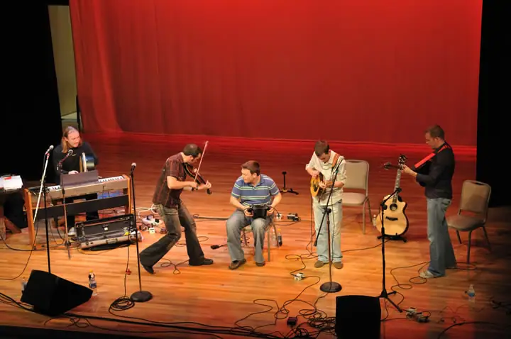 Five musicians perform on a wooden stage before a red curtain, with keyboards, bowed and stringed instruments, and microphones.