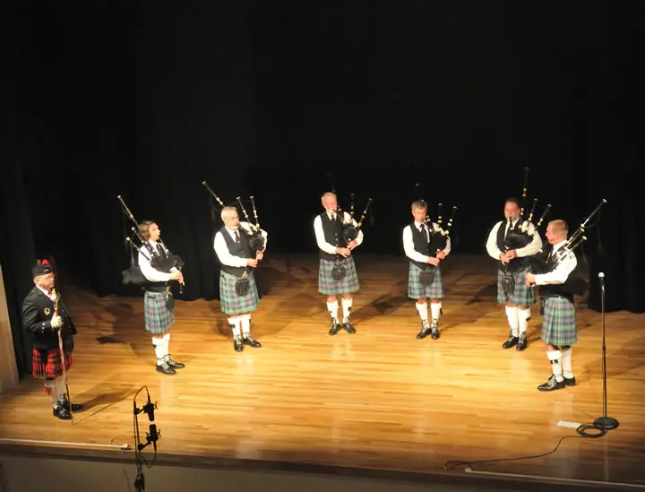 Seven kilted musicians perform with bagpipes on a wooden stage.